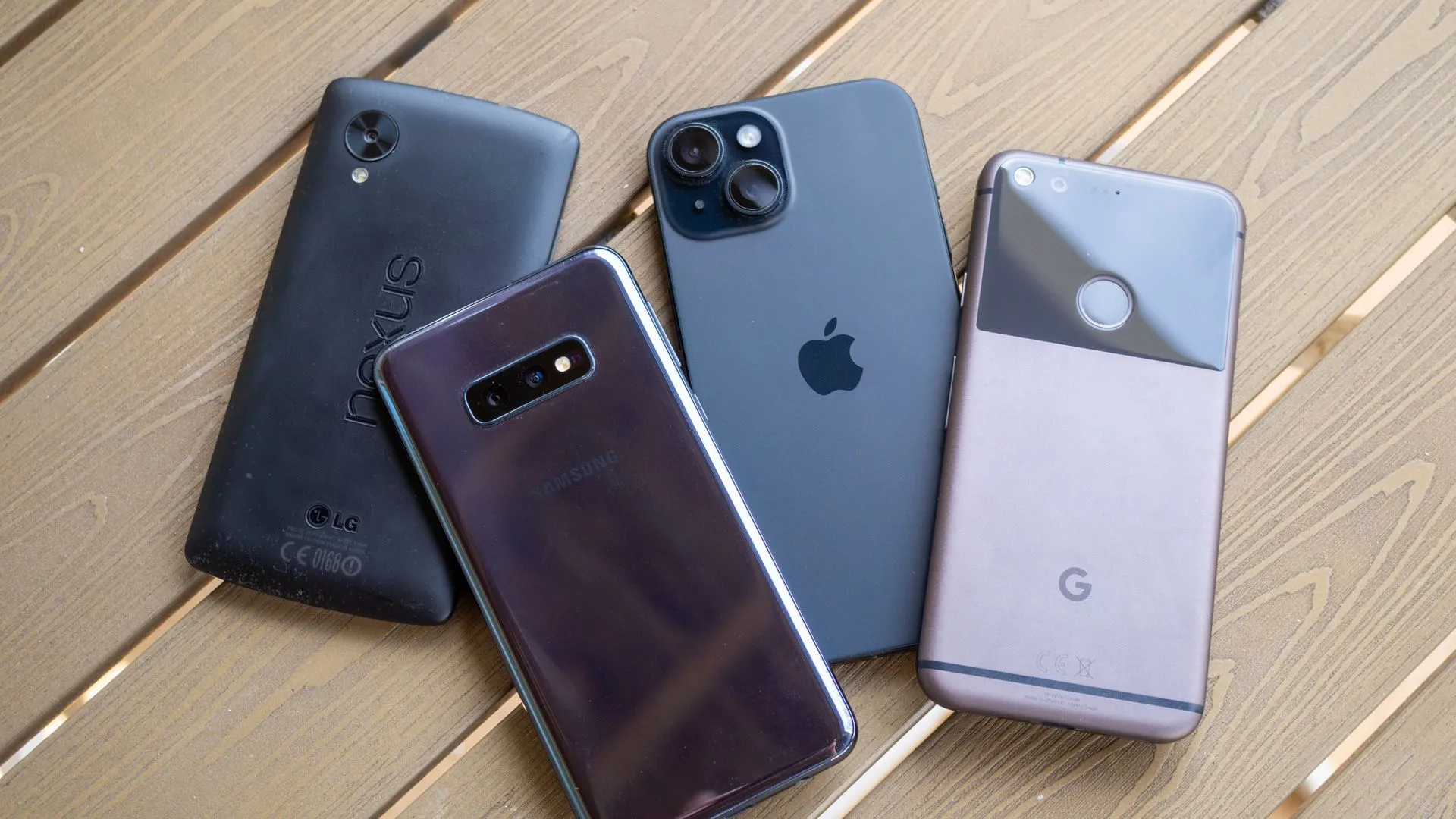 Nhiều mẫu điện thoại thông minh cũ và mới gồm Nexus 5, Galaxy S10e, iPhone 15 và Google Pixel được đặt trên mặt bàn gỗ, thể hiện sự đa dạng của các thiết bị di động.
